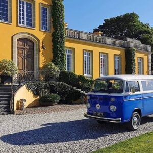 Van Volkswagen bleu/blanc stationné devant un grand bâtiment jaune avec escalier sur un chemin de gravier ensoleillé.