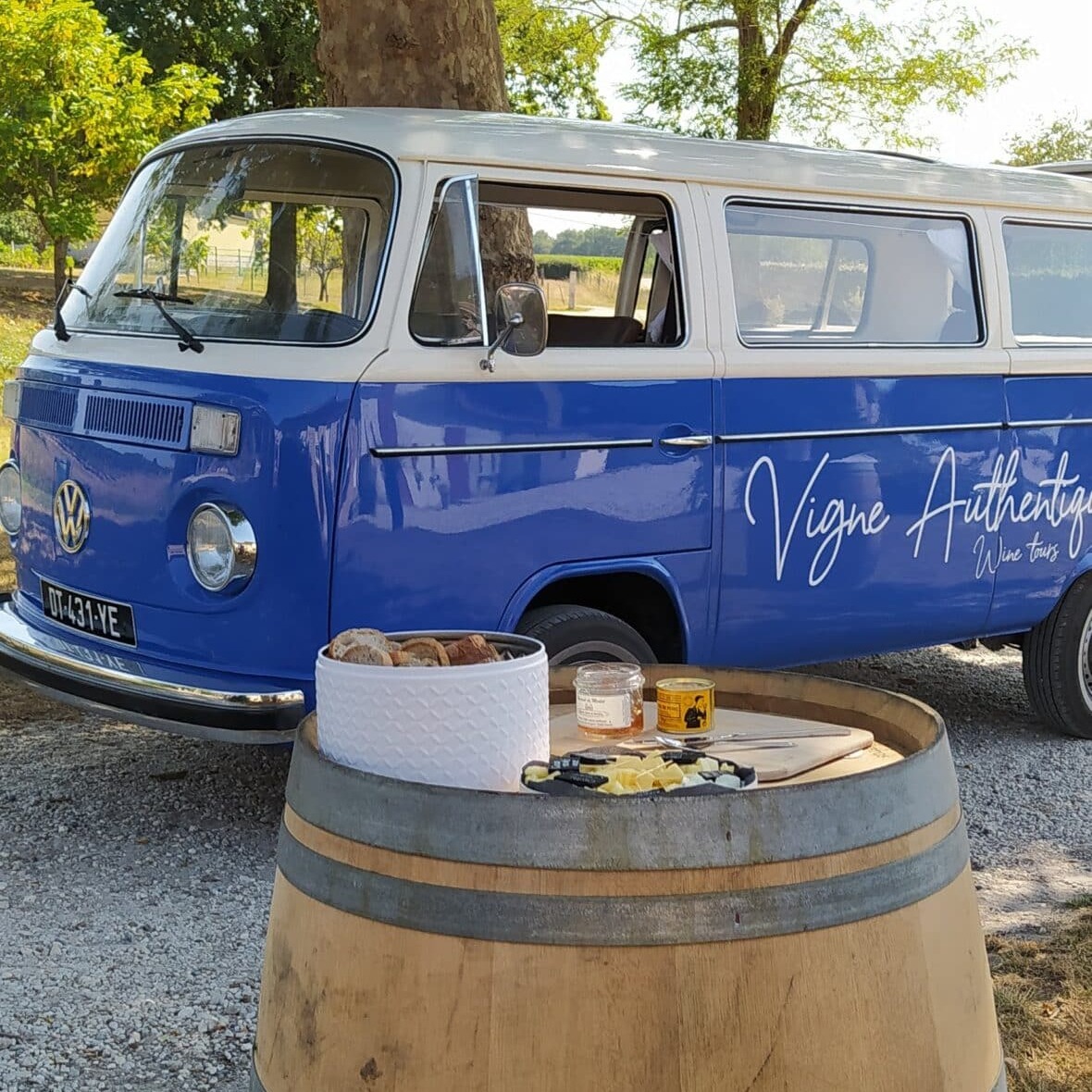VW van bleu "Vigne Authentique Wine Tours" garé à côté d'un tonneau avec pain, fromage et confiture pour dégustation.