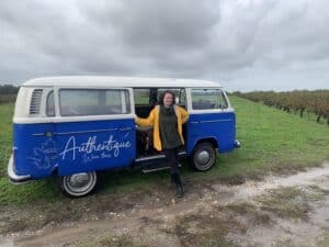 Femme souriante en imper jaune près d'un Combi VW bleu et blanc "Authentique Wine tours" dans un vignoble sous ciel gris. Chemin boueux.