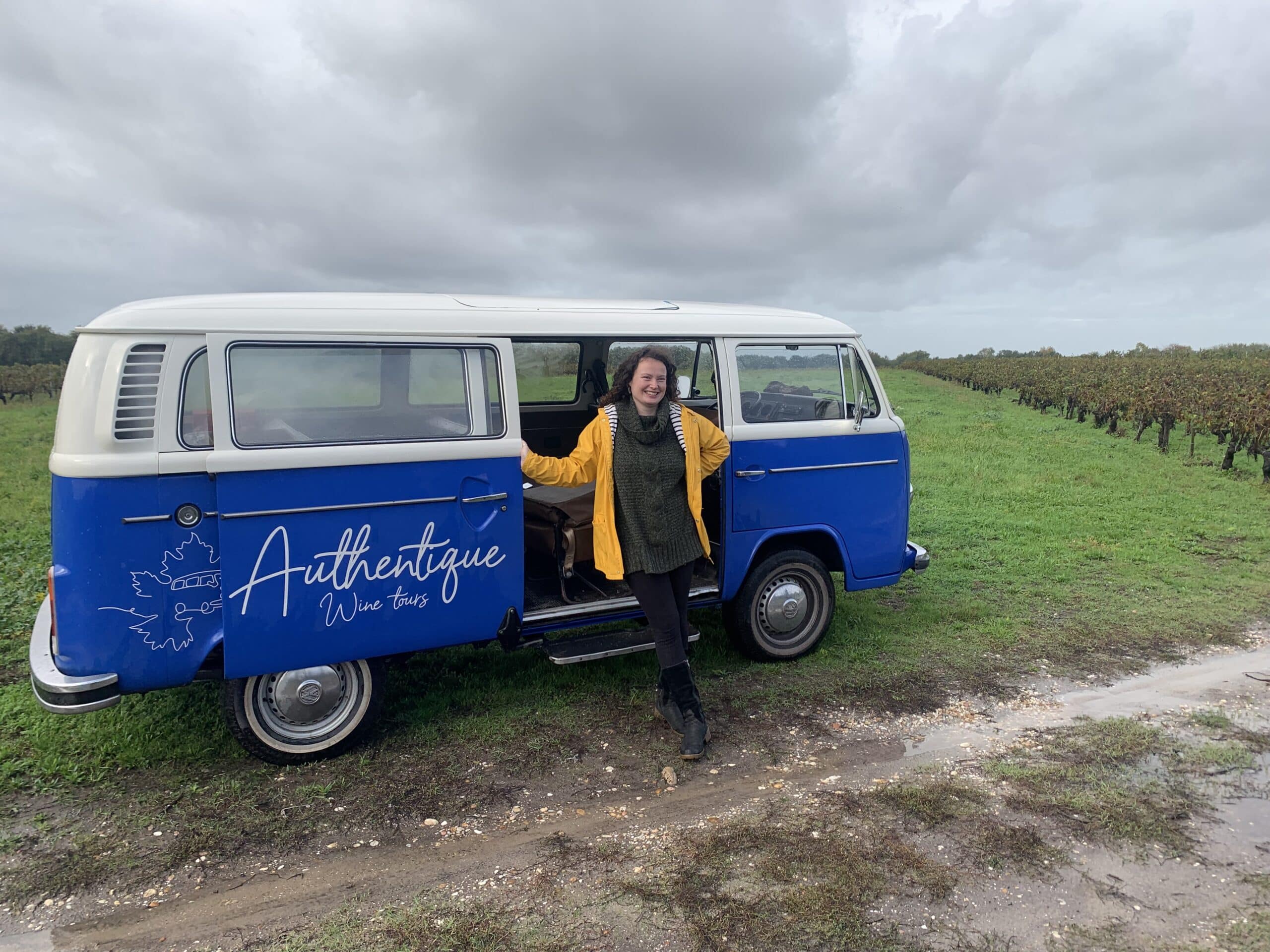 Femme souriante en imper jaune près d'un Combi VW bleu et blanc "Authentique Wine tours" dans un vignoble sous ciel gris. Chemin boueux.