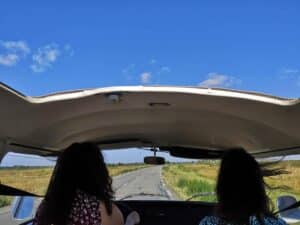 Deux femmes en van vintage, toit ouvert, sur route de campagne. Ciel bleu, champs, cheveux au vent. Une évasion libre en road trip.