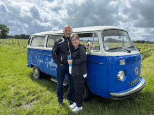 Couple souriant posant devant un Combi Volkswagen bleu et blanc, décoré de l'inscription "Vigne Authentic Wine tour", dans un vignoble par temps nuageux.