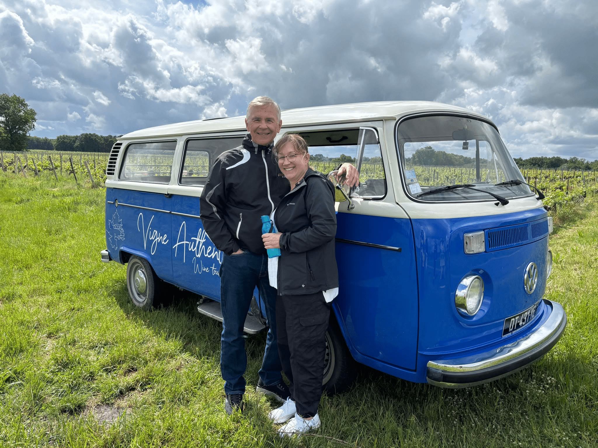 Couple souriant posant devant un Combi Volkswagen bleu et blanc, décoré de l'inscription "Vigne Authentic Wine tour", dans un vignoble par temps nuageux.