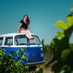 Jeune femme souriante assise sur le toit blanc et bleu d'un Combi Volkswagen immatriculé DT-431-YE, près d'une vigne.