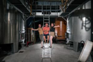 Trois personnes souriantes, deux hommes et une femme, posent sur une échelle au milieu d'une salle de cuves métalliques d'une cave.