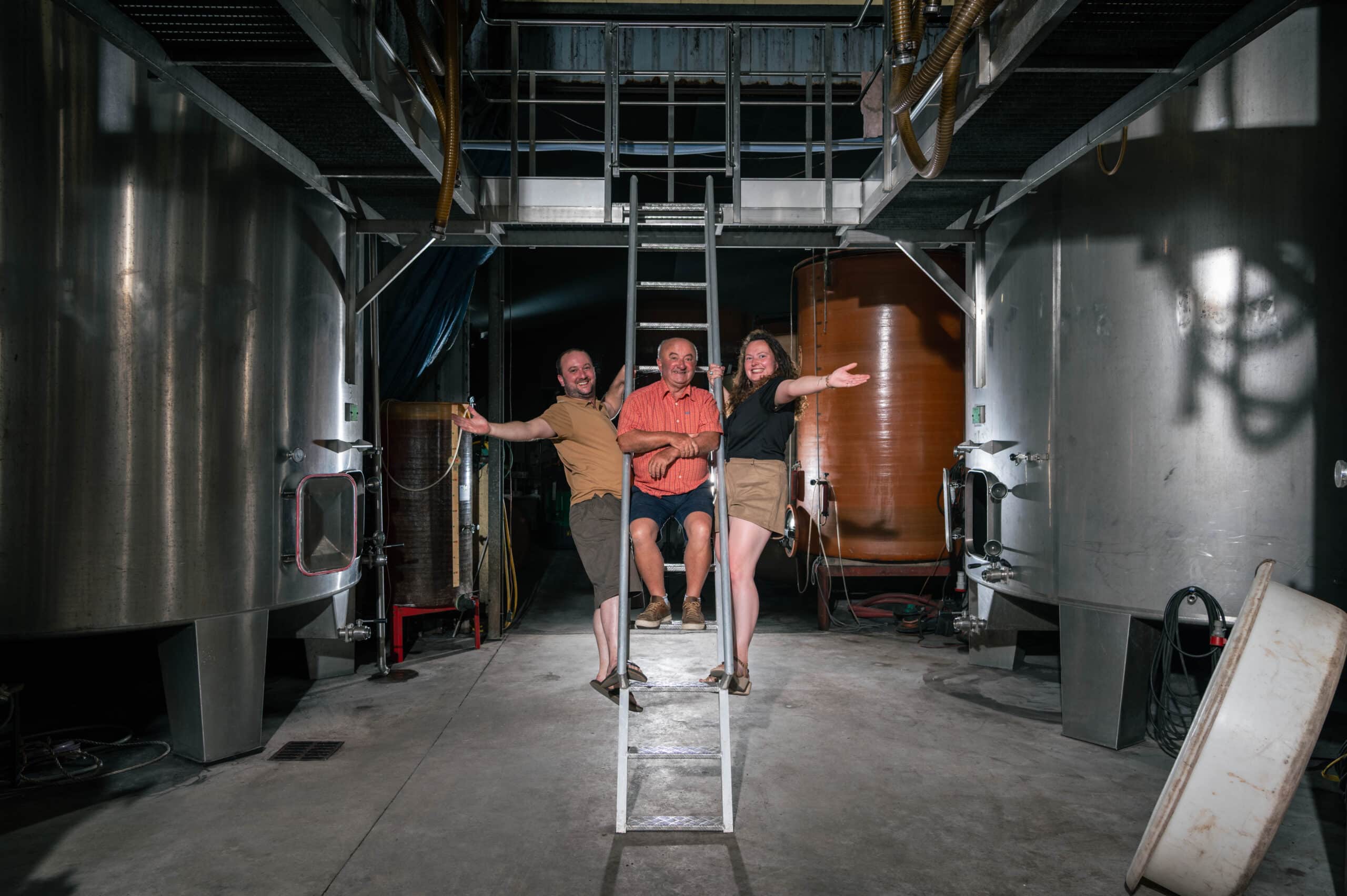 Trois personnes souriantes, deux hommes et une femme, posent sur une échelle au milieu d'une salle de cuves métalliques d'une cave.